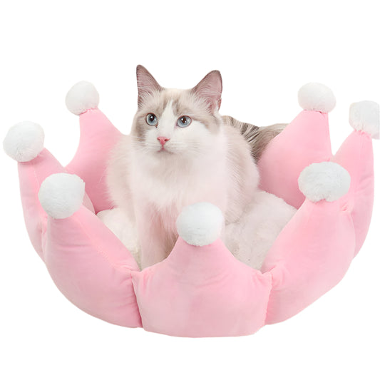 Cat sitting in a pink crown-shaped pet bed with white pom-poms on a lwhite background