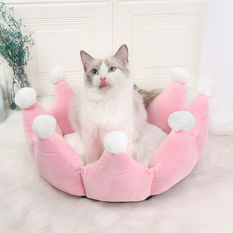 Cat sitting in a pink crown-shaped pet bed with white pom-poms on a light background
